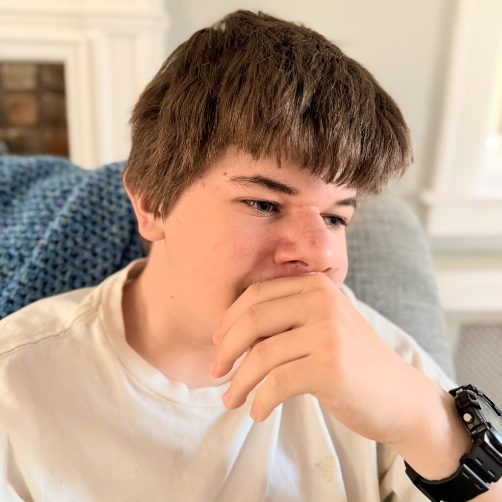 A young male with light brown hair posing with a hand over his mouth and wearing a black watch