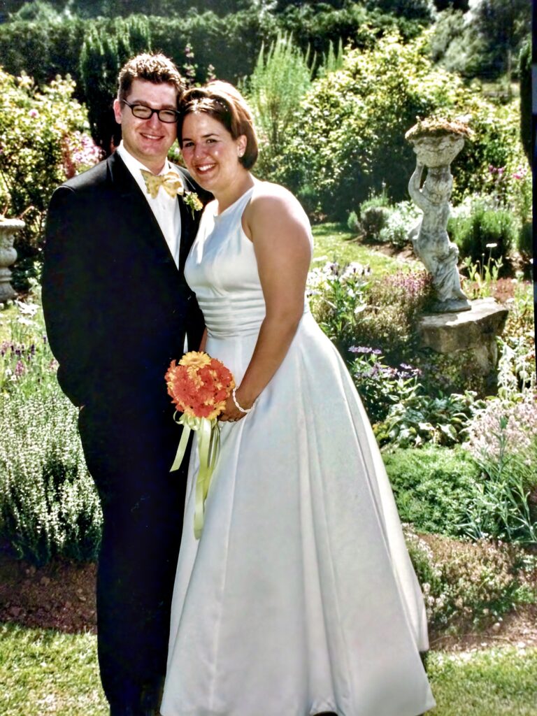 A white couple standing in a garden wearing formal wedding attire