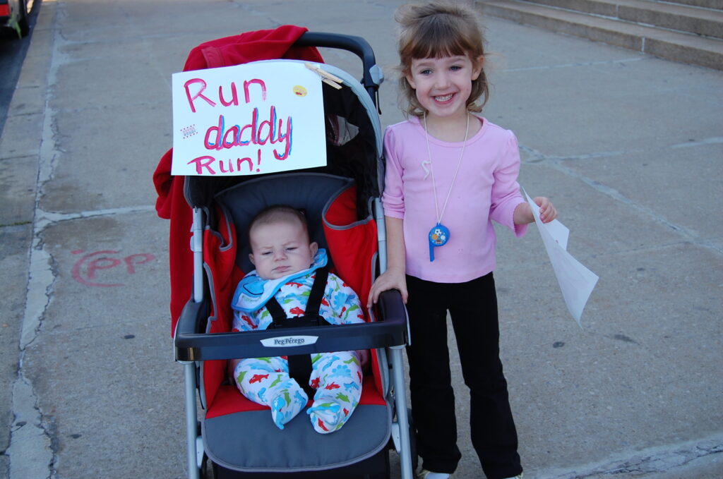 A baby in a stroller with a sign saying “Run daddy Run” next to a young girl in pink shirt smiling