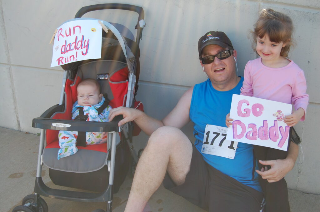 A baby in a stroller with a sign saying “Run daddy Run” next to a white male with a black hat, blue shirt and sunglasses next to a young girl in a pink shirt smiling